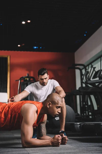 Treinador atencioso instruindo desportista afro-americano fazendo exercício prancha — Fotografia de Stock