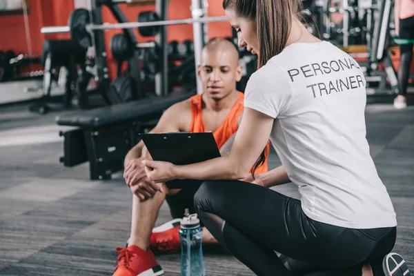 Personal trainer showing clipboard to african american sportsman sitting on floor — Stock Photo