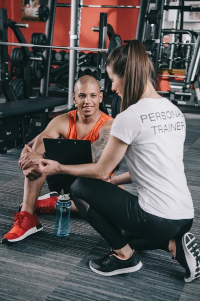 Entraîneur personnel montrant presse-papiers au sportif afro-américain souriant assis sur le sol — Photo de stock