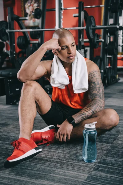 Deportista afroamericano cansado sentado en el suelo cerca de la botella de deportes - foto de stock