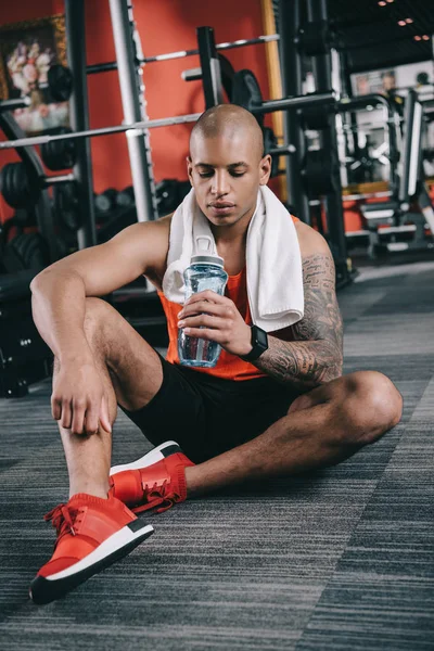 Tired african american sportsman holding sports bottle while sitting on floor — Stock Photo