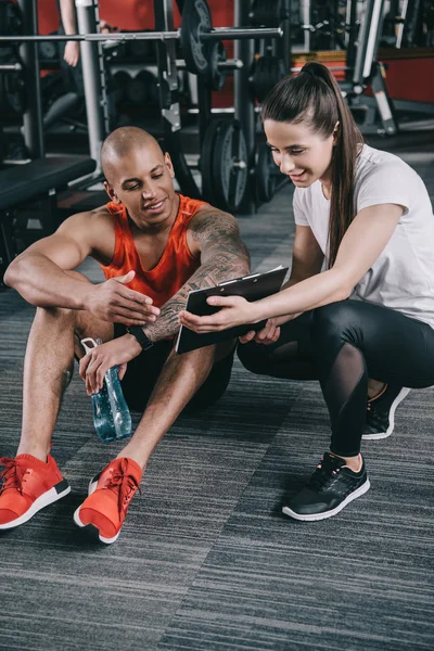 Lächelnder Trainer zeigt Klemmbrett einem fröhlichen afrikanisch-amerikanischen Sportler, der auf dem Boden sitzt — Stockfoto