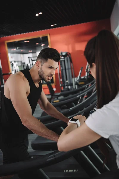 Personal trainer looking at fitness tracker while standing near sportsman running on treadmill — Stock Photo