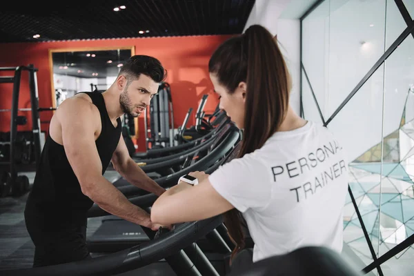 Personal trainer looking at fitness tracker while standing near sportsman running on treadmill — Stock Photo