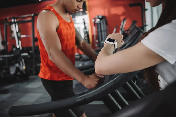 Vue recadrée de l'entraîneur regardant le tracker de remise en forme tout en se tenant près du sportif afro-américain en cours d'exécution sur tapis roulant — Photo de stock