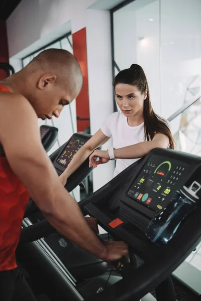 Entraîneur attentif dans le fitness tracker regardant sportif afro-américain en cours d'exécution sur tapis roulant — Photo de stock