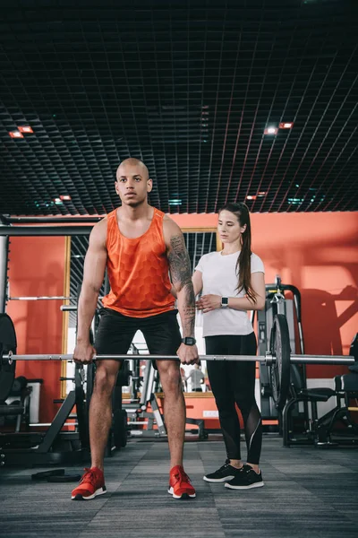 Attentive trainer standing near african american athlete lifting barbell — Stock Photo