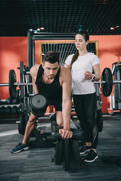 Attentive trainer assisting handsome sportsman working out with dumbbell — Stock Photo