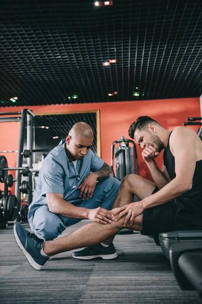 Serious african american doctor touching injured knee  of sportsman suffering from pain — Stock Photo