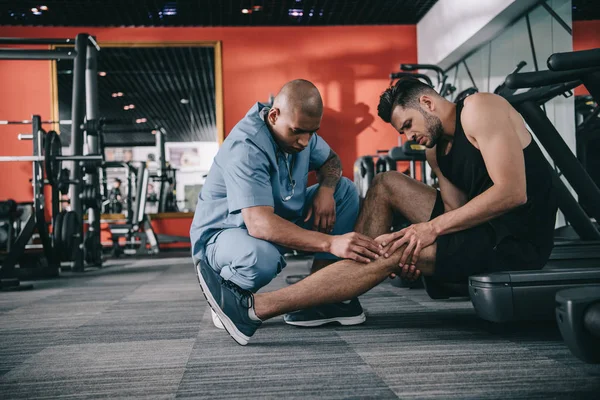 Внимательный африканский американский врач осматривает поврежденное колено спортсмена, страдающего от боли — стоковое фото