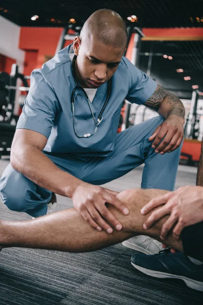 Serious african american doctor examining injured knee of sportsman — Stock Photo