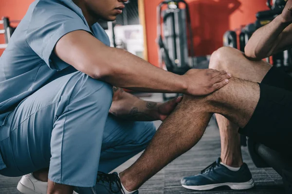 Vista recortada del médico afroamericano tocando rodilla lesionada del deportista - foto de stock