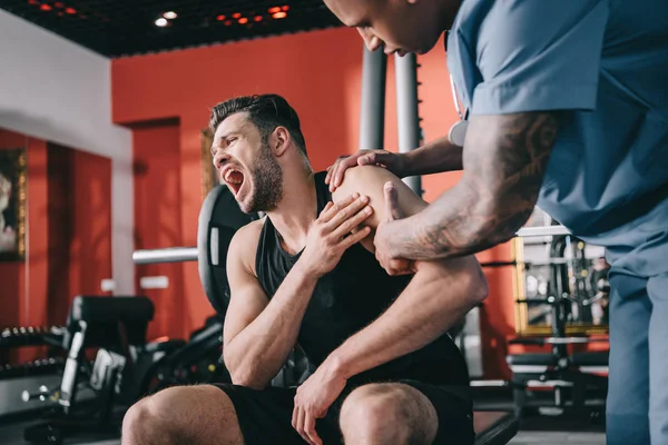 African american doctor touching injured shoulder of sreaming sportsman — Stock Photo