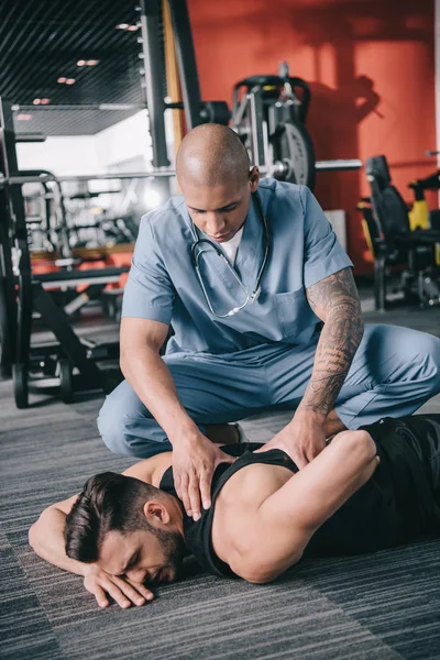 Attentive american doctor examining injured back of sportsman lying on floor — Stock Photo