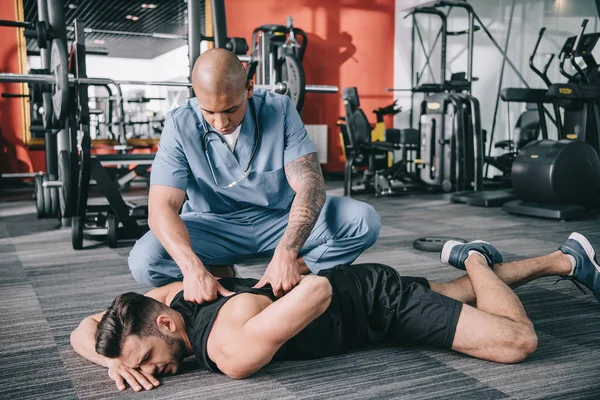 Внимательный американский врач осматривает травмированную спину лежащего на полу спортсмена — стоковое фото
