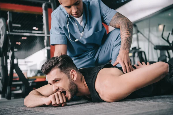 Joven médico americano tocando la espalda lesionada de deportista acostado en el suelo y sufriendo de dolor - foto de stock