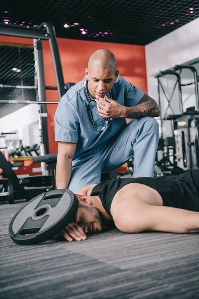 Thoughtful african american doctor near unconscious sportsman injured with weight plate — Stock Photo