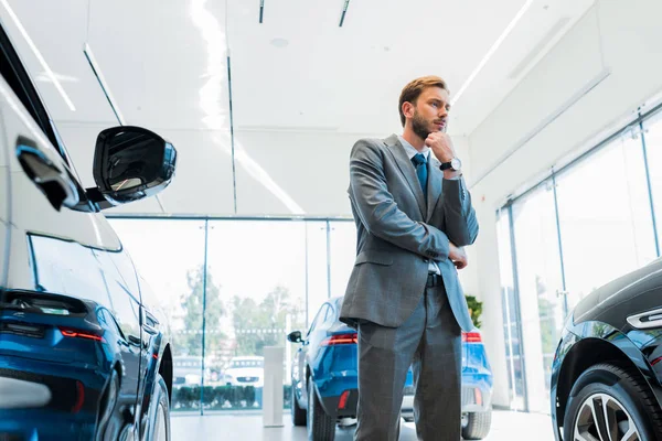 Pensive bearded businessman standing near automobiles in car showroom — Stock Photo