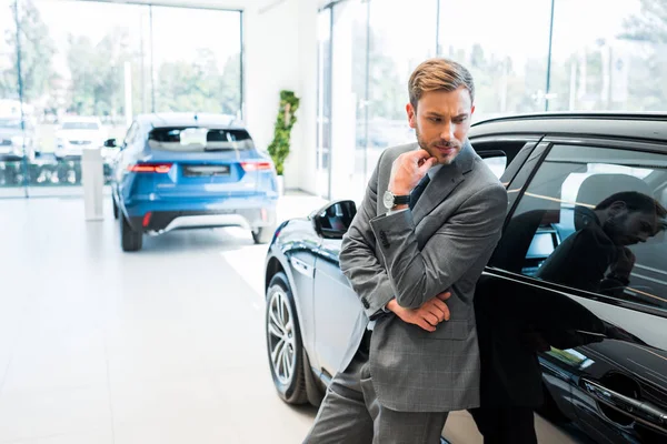 Pensativo hombre de negocios barbudo de pie cerca de los coches en la sala de exposición de coches - foto de stock