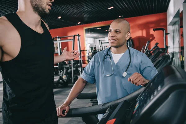 Recortado vista de deportista mostrando gesto encogiéndose de hombros cerca sonriente afroamericano médico - foto de stock