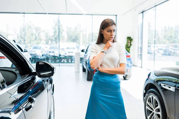 Giovane e pensierosa donna in piedi vicino alle auto in auto showroom — Foto stock