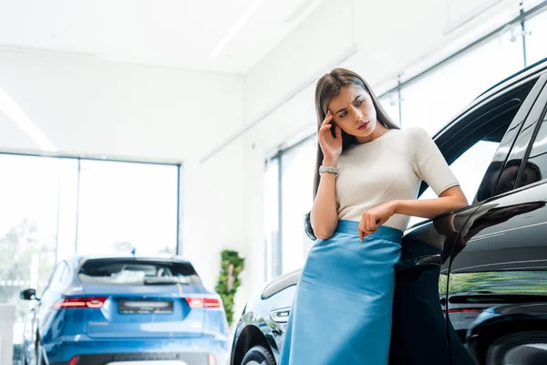 Attraktive und nachdenkliche Frau steht in Autosalon neben Autos — Stockfoto