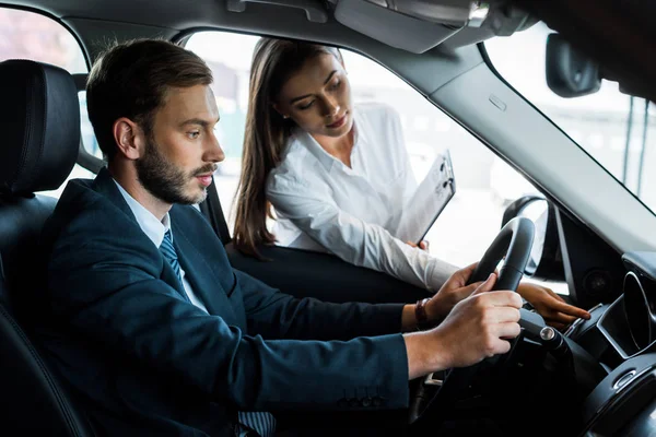 Atractiva mujer sujetando el portapapeles y apuntando con el dedo al botón cerca del volante - foto de stock