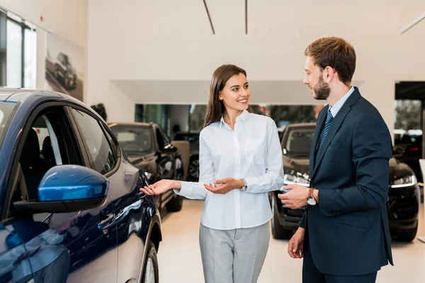 Positive car dealer gesturing while looking at bearded man in car showroom — Stock Photo