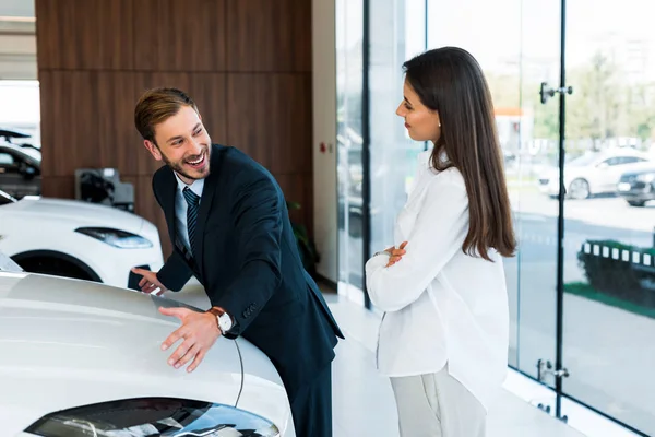 Heureux concessionnaire de voiture barbu geste près de femme attrayante debout avec les bras croisés dans la salle d'exposition de voiture — Photo de stock