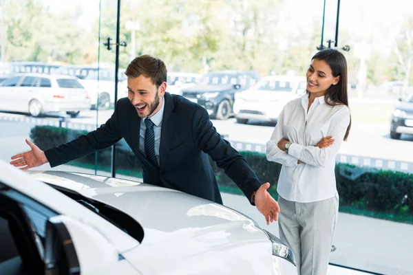 Foco seletivo de revendedor de carro barbudo feliz gesticulando perto de mulher bonita de pé com braços cruzados no showroom carro — Fotografia de Stock