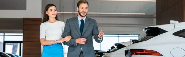 Tiro panorâmico de homem de terno gesticulando perto de mulher bonita no showroom do carro — Fotografia de Stock