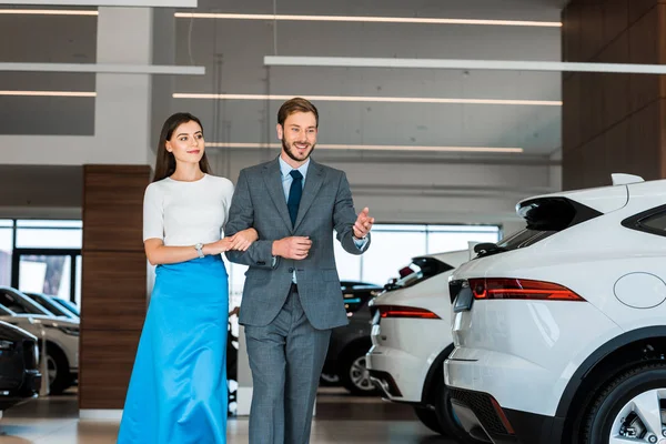 Happy bearded man gesturing while walking with woman in car showroom — Stock Photo