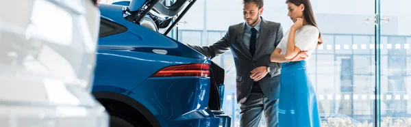 Panoramic shot of attractive woman and handsome man looking at car trunk in car showroom — Stock Photo