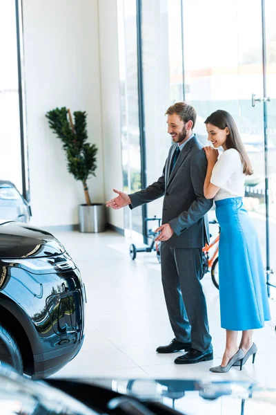 Selektiver Fokus einer fröhlichen Frau, die mit einem hübschen bärtigen Mann steht und gestikuliert, während sie das Auto betrachtet — Stockfoto