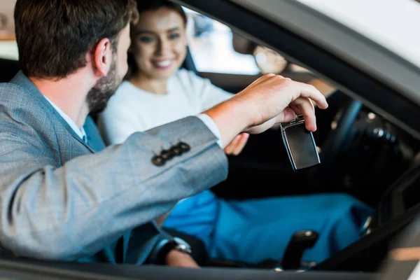 Селективное внимание бородатого мужчины, держащего ключ от машины рядом с улыбающейся женщиной, сидящей в машине — стоковое фото