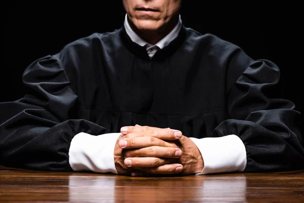 Cropped view of judge in judicial robe sitting at table with clenched hands isolated on black — Stock Photo