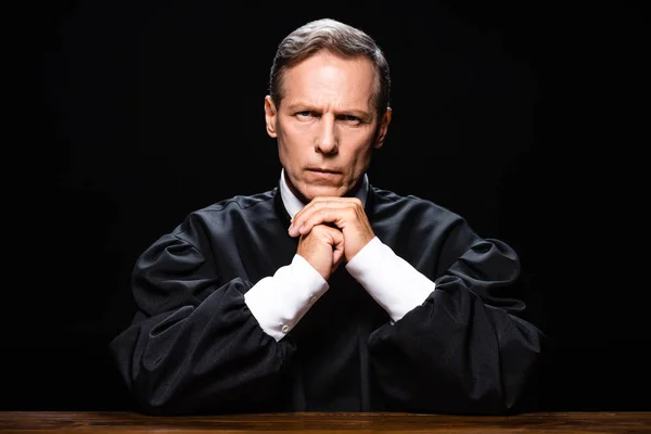Judge in judicial robe sitting at table and looking at camera isolated on black — Stock Photo