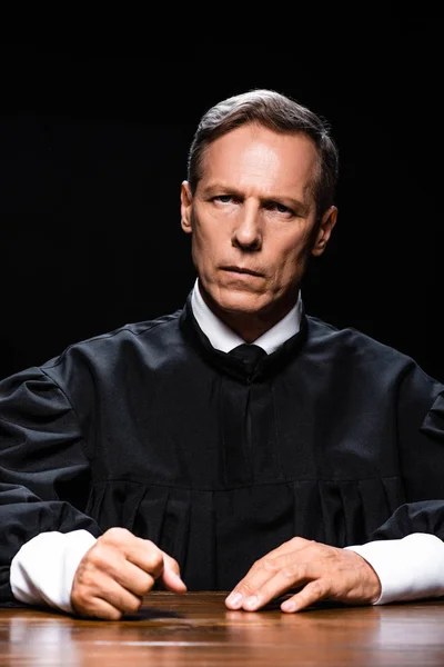 Judge in judicial robe sitting at table and looking at camera isolated on black — Stock Photo