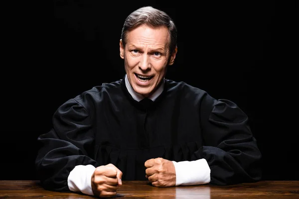 Judge in judicial robe sitting at table and looking at camera and screaming isolated on black — Stock Photo