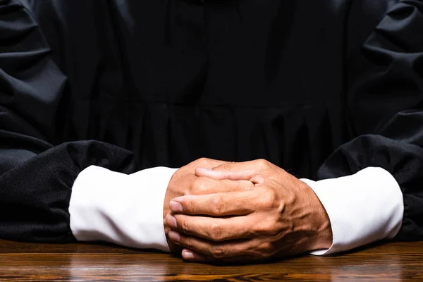 Vista recortada del juez en bata judicial sentado a la mesa con las manos apretadas aisladas en negro - foto de stock