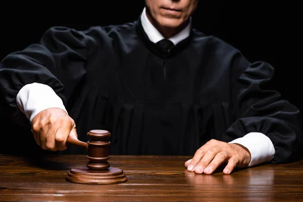 Vista recortada del juez en bata judicial sentado en la mesa y golpeando con mazo aislado en negro - foto de stock