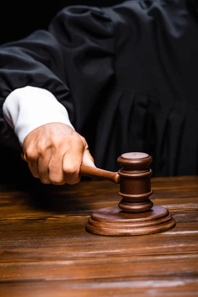 Cropped view of judge in judicial robe sitting at table and hitting with gavel isolated on black — Stock Photo