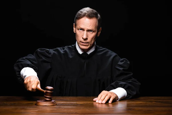 Judge in judicial robe sitting at table and hitting with gavel isolated on black — Stock Photo