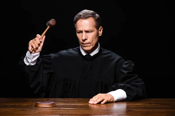 Суддя в судовому халаті, сидячи за столом і вдарившись з даром ізольовано на чорному — стокове фото