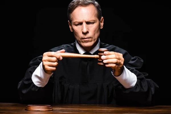 Judge in judicial robe sitting at table and holding gavel isolated on black — Stock Photo