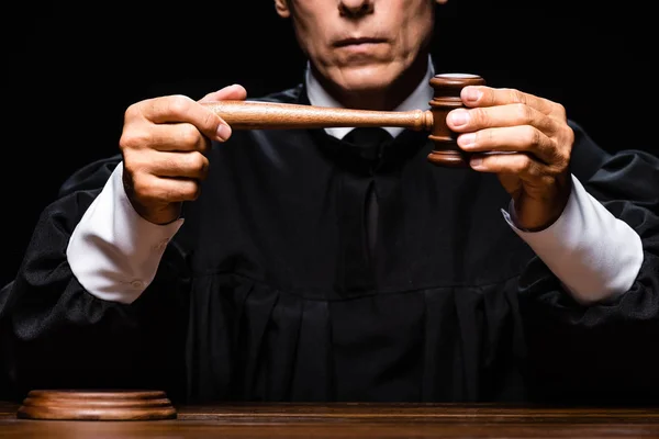 Cropped view of judge in judicial robe sitting at table and holding gavel isolated on black — Stock Photo