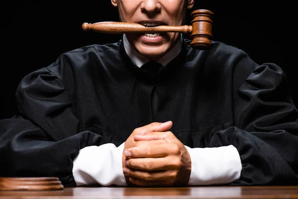 Cropped view of judge in judicial robe sitting at table and holding gavel in mouth isolated on black — Stock Photo