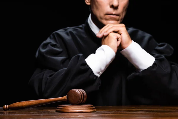 Vista recortada del juez en bata judicial sentado a la mesa con mazo aislado en negro - foto de stock