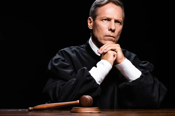Judge in judicial robe sitting at table with gavel isolated on black — Stock Photo
