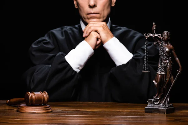 Cropped view of judge in judicial robe sitting at table with gavel and themis figure isolated on black — Stock Photo